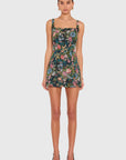 A model wearing a sleeveless mini dress with a dark green floral print, featuring a square neckline and thin shoulder straps, paired with black high heels against a light gray background.