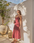 Woman in a red polka dot dress standing against a white wall with a plant and pot in the background.
