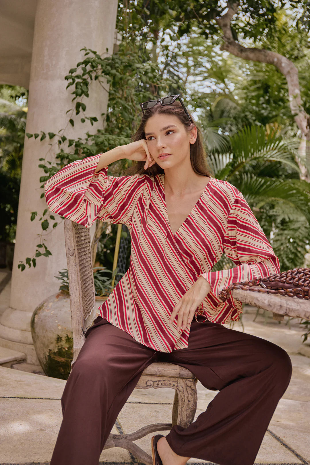 Woman in a striped red and white blouse and brown pants sitting outdoors.