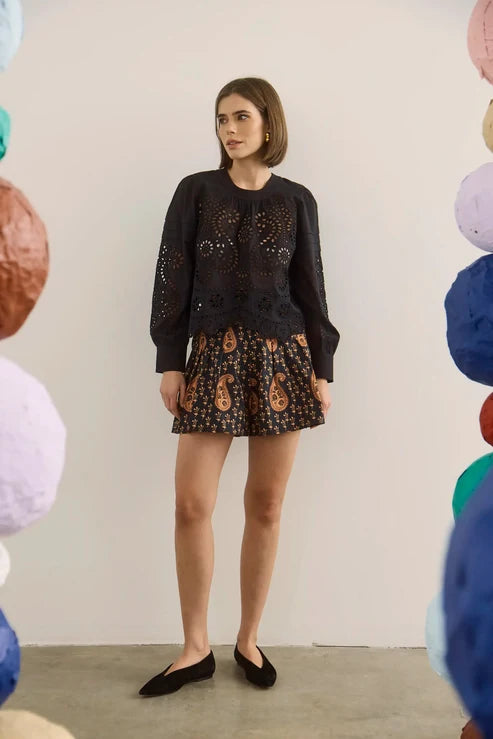 A woman stands in a studio wearing a black long-sleeve lace-trim top and a black shorts with a brown paisley pattern. She is framed by colorful hanging paper lanterns on both sides.