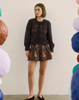 A woman stands in a studio wearing a black long-sleeve lace-trim top and a black shorts with a brown paisley pattern. She is framed by colorful hanging paper lanterns on both sides.