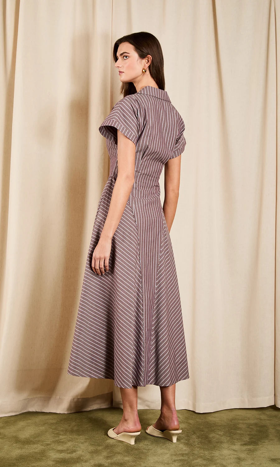 A woman modeling a short-sleeve midi dress with vertical purple and beige stripes, shown from a side-back angle against a draped beige curtain backdrop.
