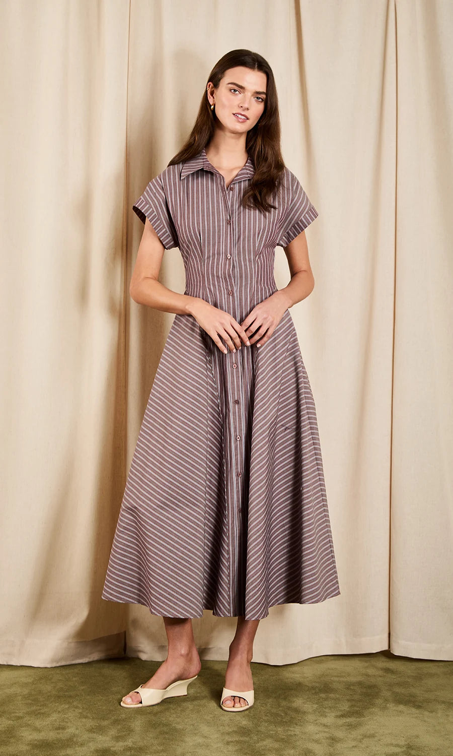A woman modeling a short-sleeve button-front midi dress with vertical stripes in muted purple and beige, standing against a draped beige curtain backdrop.