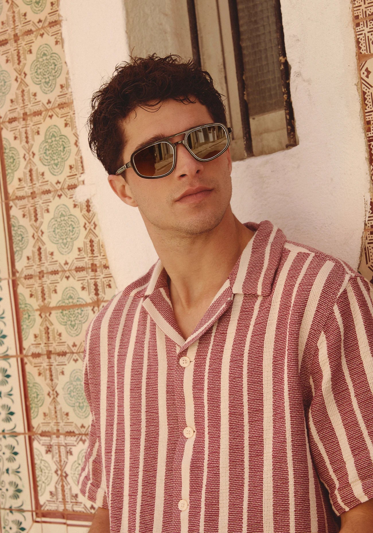 Man wearing sunglasses and a striped shirt against a patterned wall.