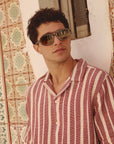 Man wearing sunglasses and a striped shirt against a patterned wall.