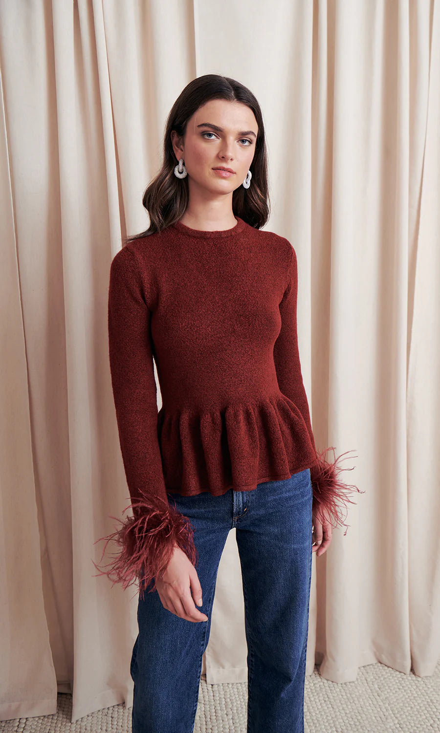 Woman wearing a rust-colored peplum sweater and blue jeans against a beige curtain background