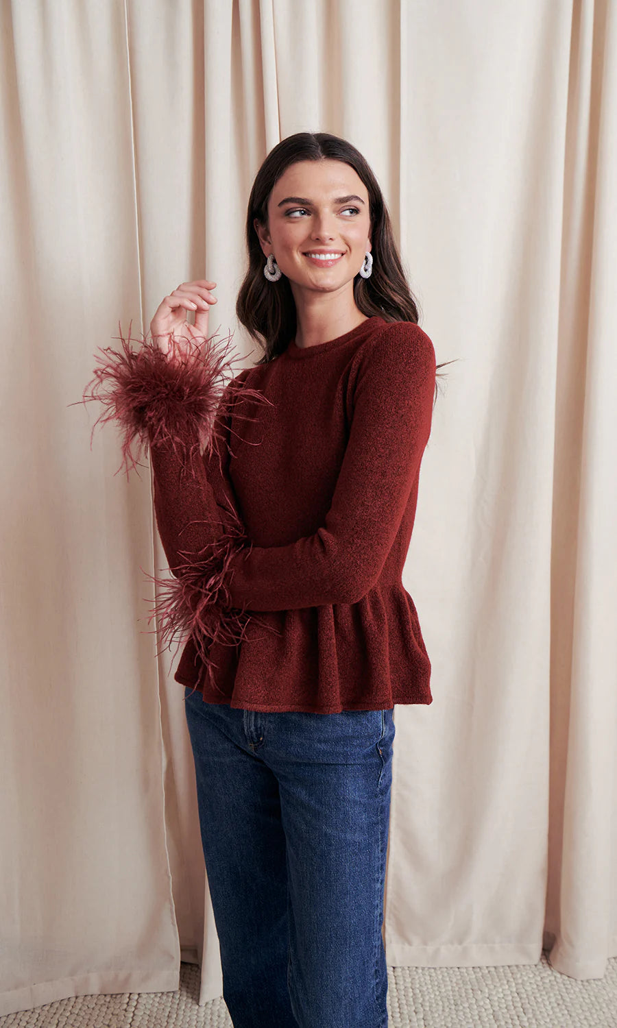 Woman wearing a burgundy sweater with feather details and blue jeans against a beige curtain background, side/front view