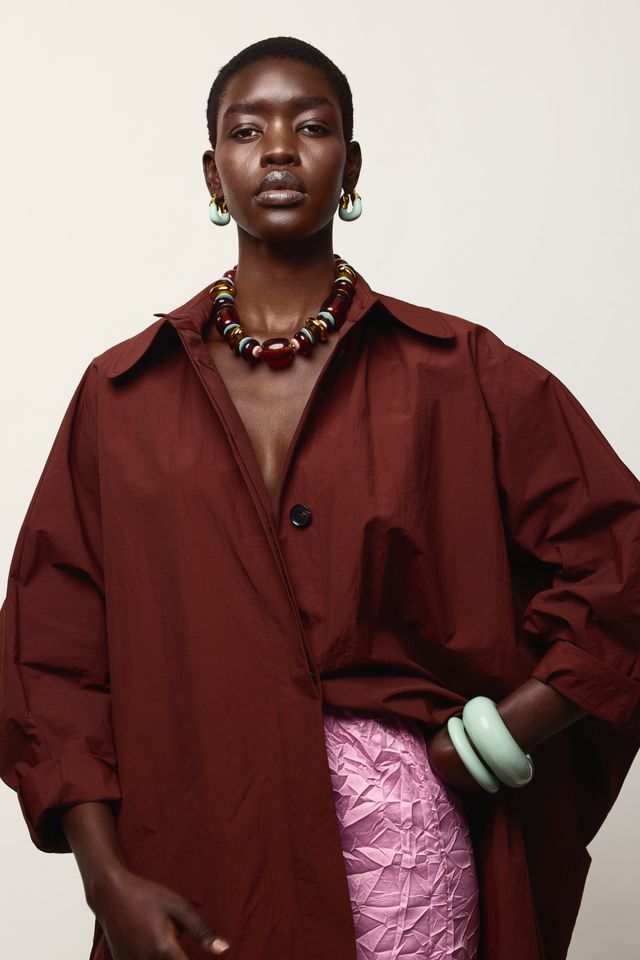 A model wearing an oversized dark red button-up shirt with a pink textured skirt, a chunky beaded necklace, and light green earrings, set against a plain off-white background.