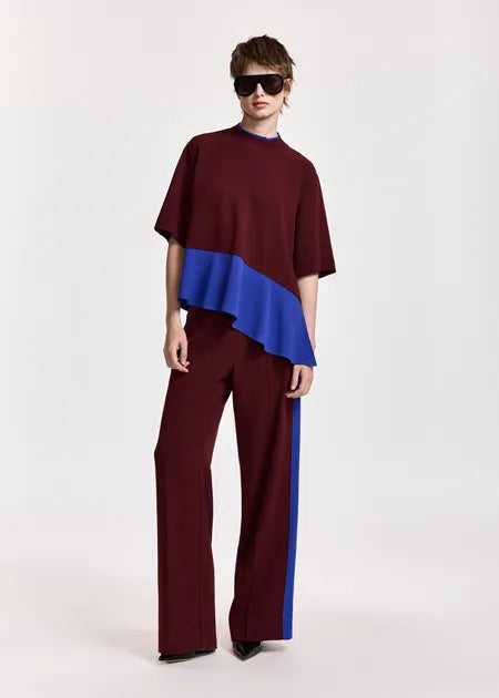A model wearing a color-blocked two-piece outfit consisting of a short-sleeve top and matching wide-leg trousers. The top is burgundy with a bright blue ruffled overlay, and the trousers are burgundy with a matching blue side stripe.
