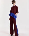 A model wearing a two-piece color-blocked outfit consisting of a high-neck, short-sleeved top with a peplum hem in burgundy and royal blue, paired with matching wide-leg trousers with blue side panels. The model is also wearing black sunglasses.
