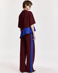 A model is shown from the back wearing a two-piece outfit consisting of a burgundy top with a bright blue ruffled underlayer and matching wide-leg trousers with a blue side stripe, set against a plain light gray background.