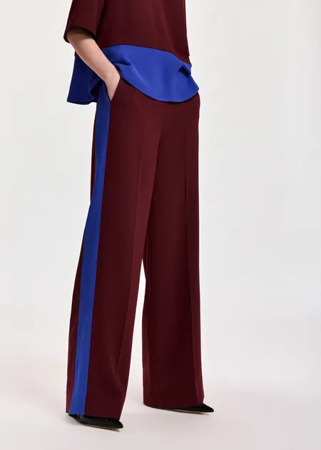 A model wearing wide-leg trousers in deep burgundy with a bold royal blue side stripe, paired with a matching top and black pointed shoes, against a plain light background.