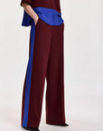 A model wearing wide-leg trousers in deep burgundy with a bold royal blue side stripe, paired with a matching top and black pointed shoes, against a plain light background.
