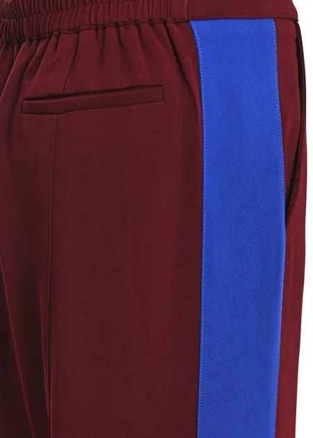 Close-up of a pair of track pants showing the side pocket, elastic waistband, and a bold vertical royal blue stripe on a burgundy fabric background.
