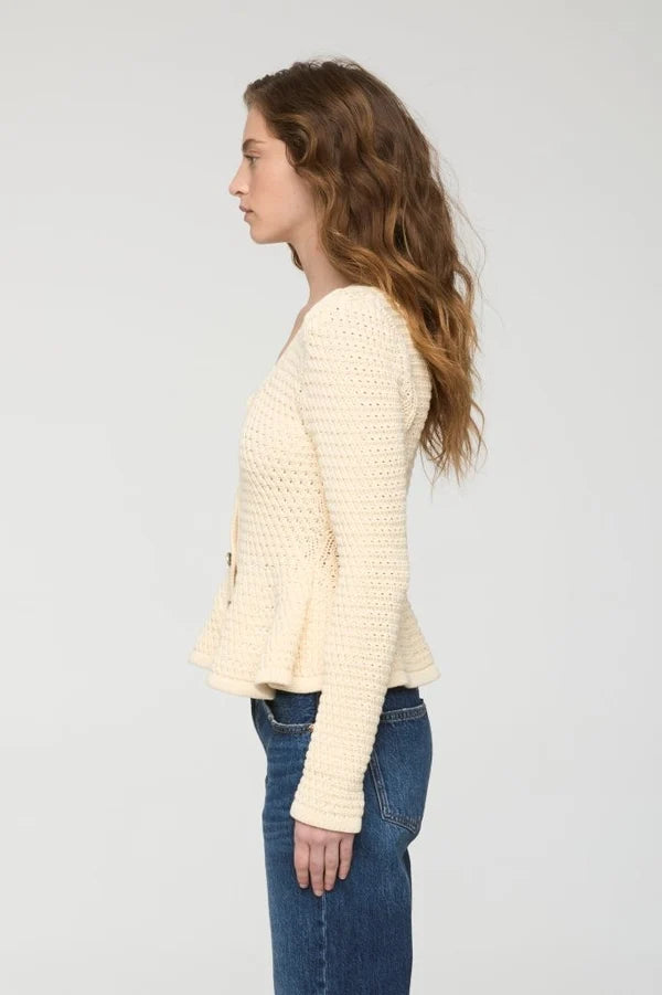 A woman shown in profile wearing a cream-colored, textured knit cardigan with a peplum hem and blue jeans, set against a plain light gray studio background.