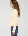 A woman shown in profile wearing a cream-colored, textured knit cardigan with a peplum hem and blue jeans, set against a plain light gray studio background.