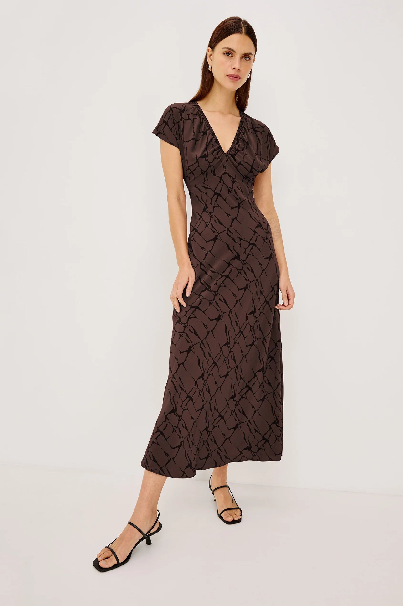 A woman modeling a dark brown midi wrap dress with a textured abstract pattern, short flutter sleeves, and a V-neckline, paired with black strappy sandals against a plain white studio background.