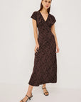 A woman modeling a dark brown midi wrap dress with a textured abstract pattern, short flutter sleeves, and a V-neckline, paired with black strappy sandals against a plain white studio background.