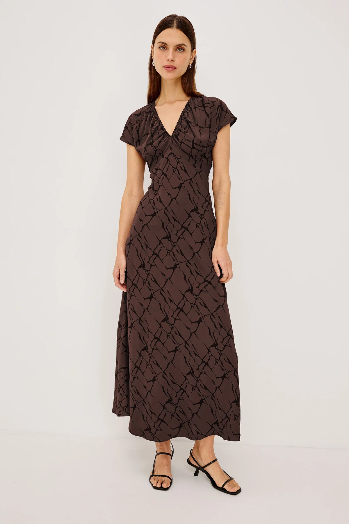 A woman modeling a dark brown midi wrap dress with a textured abstract pattern, short flutter sleeves, and a V-neckline, paired with black strappy sandals against a plain white studio background.