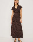 A woman modeling a dark brown midi wrap dress with a textured abstract pattern, short flutter sleeves, and a V-neckline, paired with black strappy sandals against a plain white studio background.