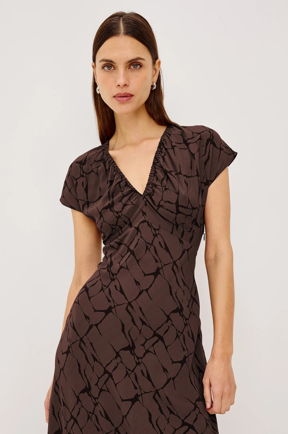 A woman modeling a dark brown midi wrap dress with a textured abstract pattern, short flutter sleeves, and a V-neckline, against a plain white studio background.