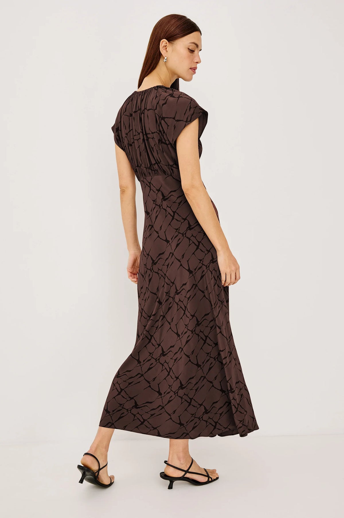 A woman modeling a dark brown midi dress with a black abstract pattern, shown from a rear three-quarter angle against a plain white studio background.