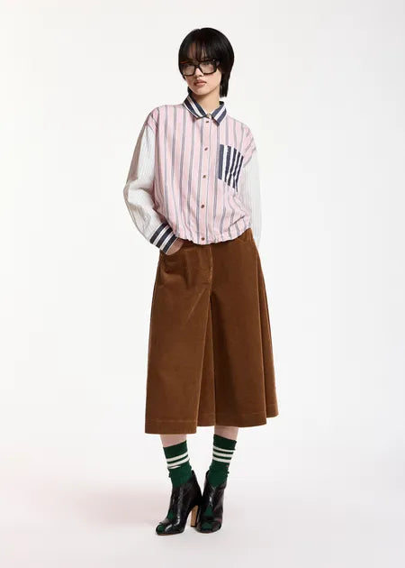 A model stands against a plain white studio background wearing a pink and white vertically striped button-up shirt with contrasting white sleeves, a brown wide-leg midi skirt, green striped socks, and black high-heeled boots.