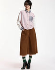 A model stands against a plain white studio background wearing a pink and white vertically striped button-up shirt with contrasting white sleeves, a brown wide-leg midi skirt, green striped socks, and black high-heeled boots.