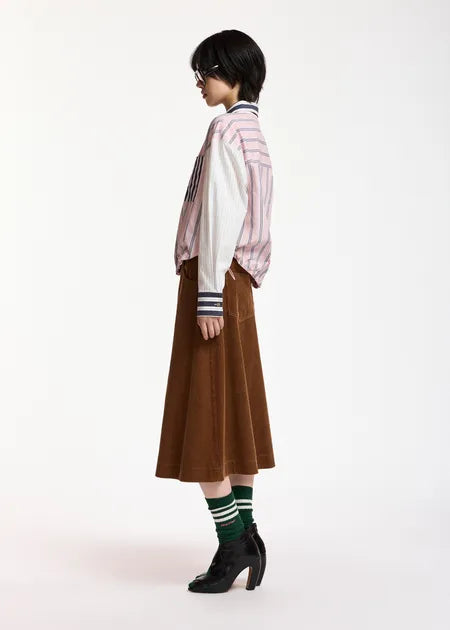 A model is shown from a side profile wearing a pink and white striped button-up shirt, a brown midi skirt, green and white striped socks, and black ankle boots, set against a plain white studio background.