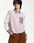 A model wearing a pink and white vertically striped button-up shirt with navy blue striped accents on the collar, cuffs, and chest pocket, paired with brown corduroy pants and black sunglasses, against a plain white background.
