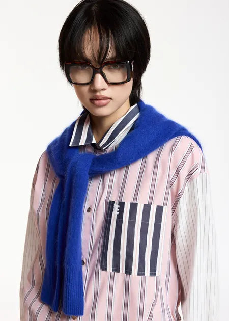 A model wearing a pink and white vertically striped button-up shirt with a striped chest pocket, a vibrant blue fuzzy knit scarf, and black-rimmed glasses, set against a plain white studio background.