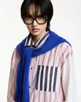 A model wearing a pink and white vertically striped button-up shirt with a striped chest pocket, a vibrant blue fuzzy knit scarf, and black-rimmed glasses, set against a plain white studio background.