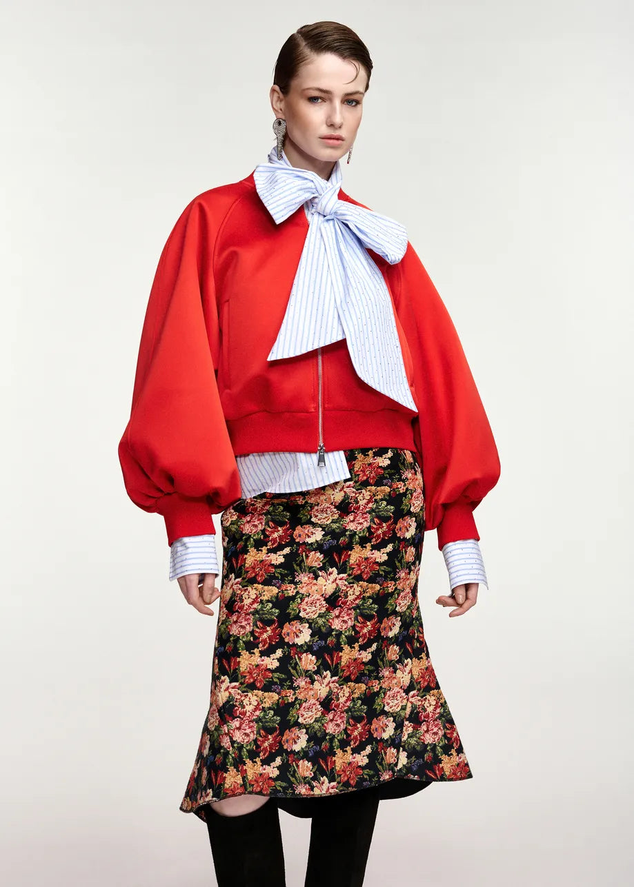 A model wearing a vibrant red cropped jacket with dramatic balloon sleeves, a large light blue and white striped bow tie, and a black midi skirt with a dense red, pink, and orange floral print.