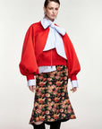 A model wearing a vibrant red cropped jacket with dramatic balloon sleeves, a large light blue and white striped bow tie, and a black midi skirt with a dense red, pink, and orange floral print.