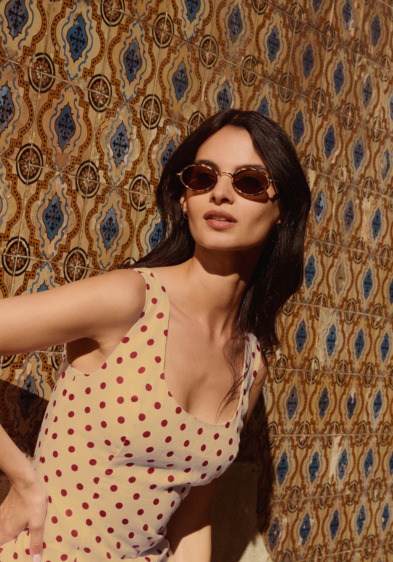 Woman wearing a polka dot dress and sunglasses against a patterned wall.