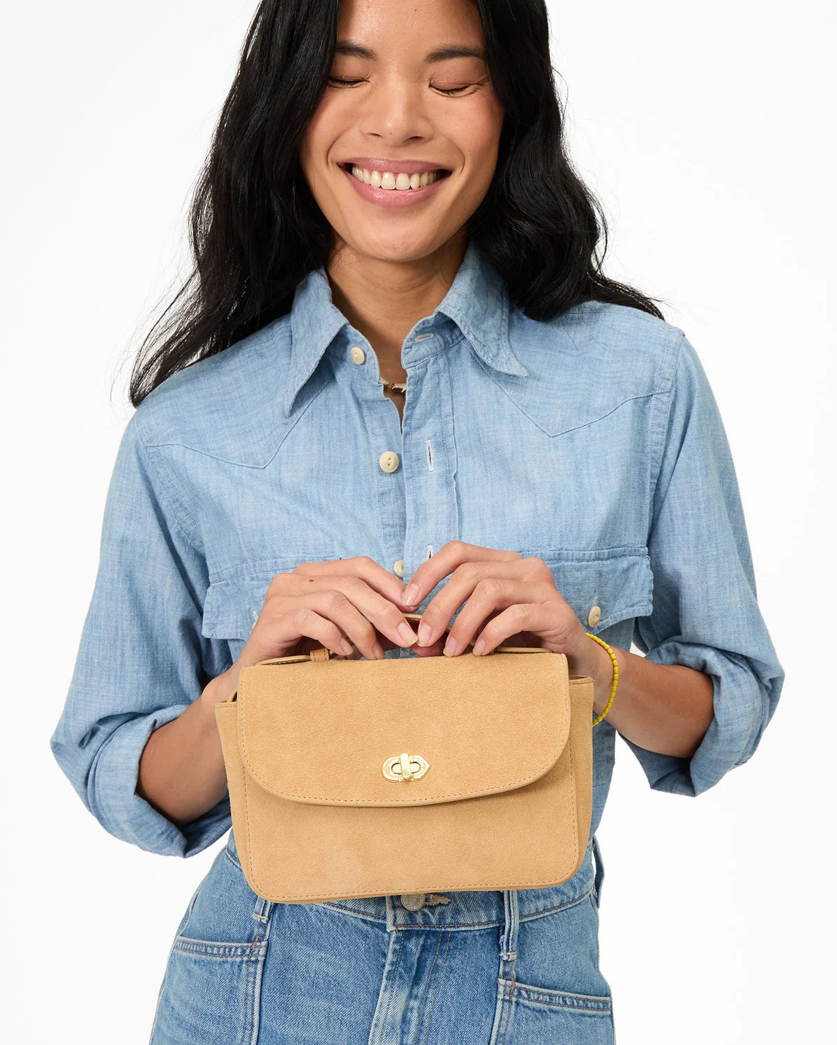 Woman holding the top handle of a  tan crossbody bag against a white background