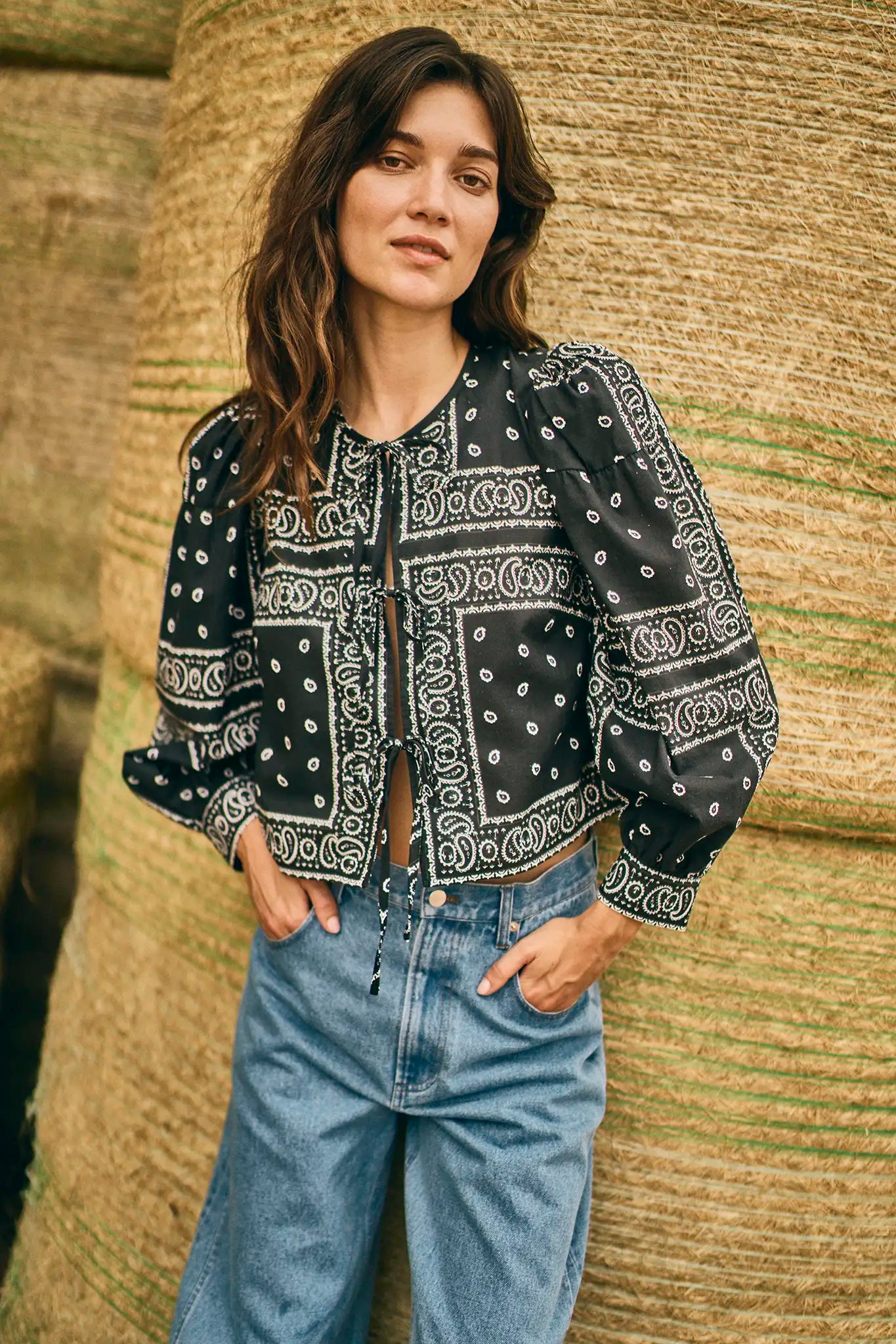 Woman wearing a black patterned blouse and blue jeans standing against hay bails.