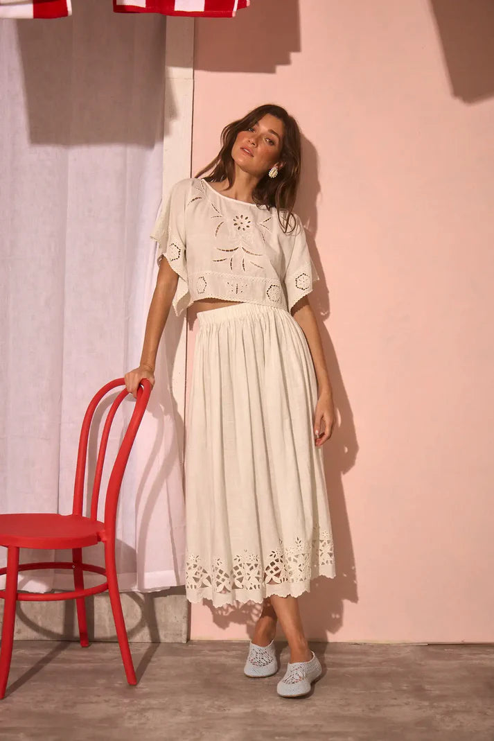 Woman in a white outfit standing next to a red chair against a pink wall.