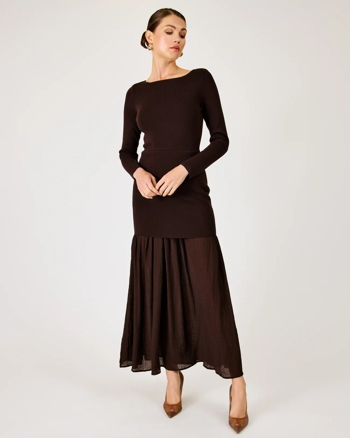 A woman modeling a dark brown long-sleeve maxi dress with a fitted bodice and a flowing semi-sheer skirt, paired with brown pointed-toe heels, against a plain light gray studio background.