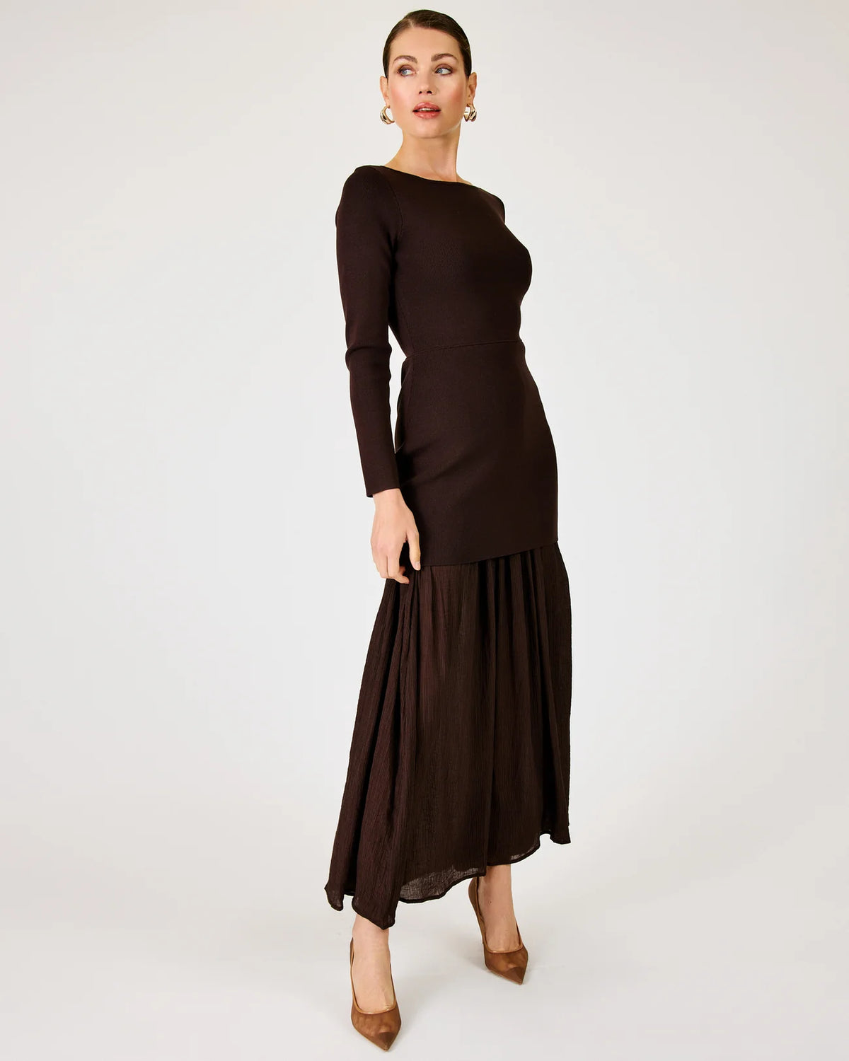 A woman modeling a dark brown long-sleeve maxi dress with a boat neckline and a pleated skirt, standing against a plain light gray studio background.