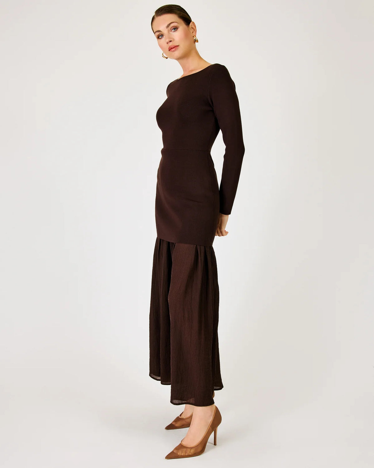 A woman modeling a dark brown long-sleeve maxi dress with a boat neckline and a pleated, flowing skirt, paired with brown pointed-toe heels against a light gray studio background.