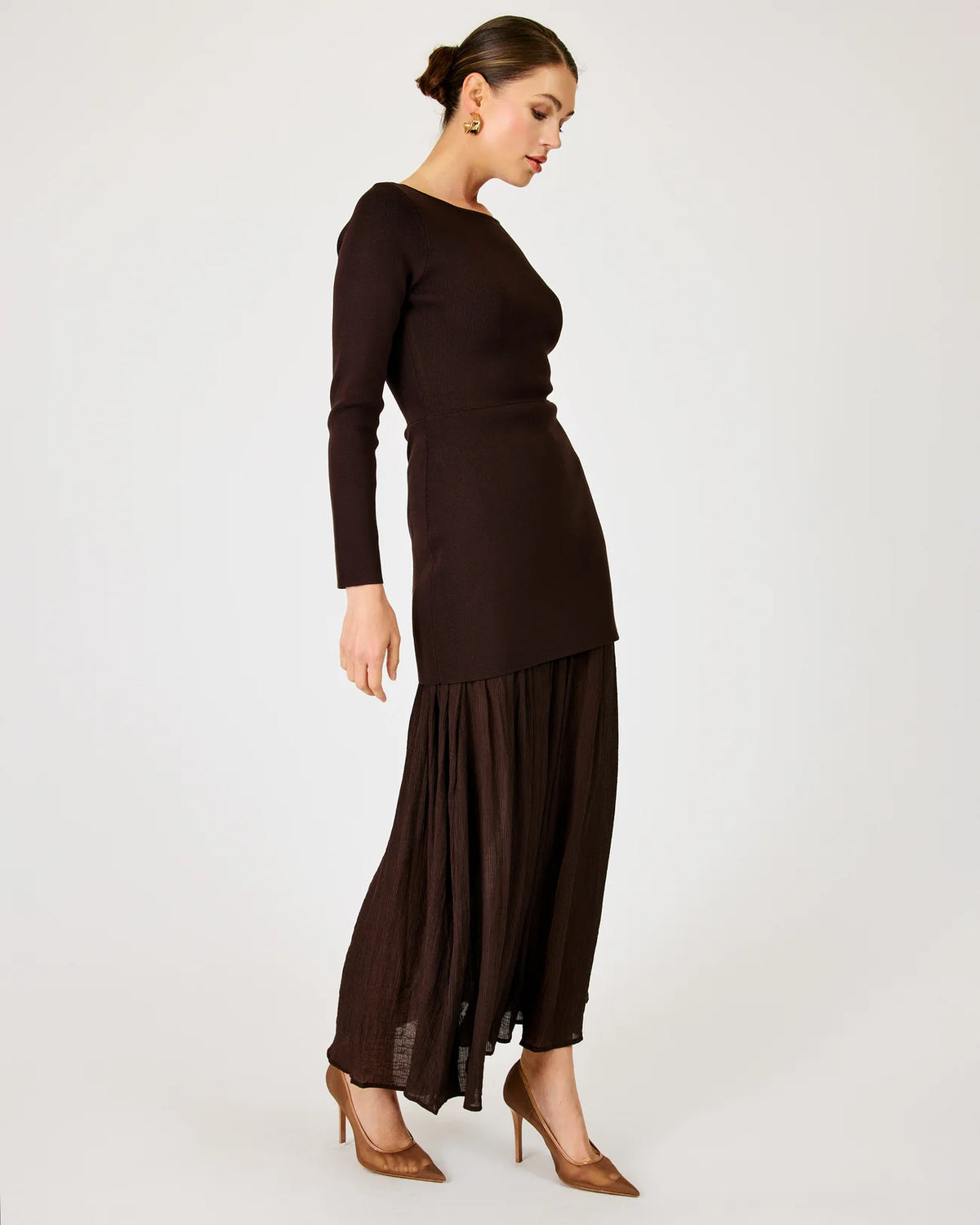 A woman modeling a dark brown ribbed knit long-sleeve maxi dress with a pleated, flowing skirt, paired with brown pointed-toe heels, against a plain light gray studio background.