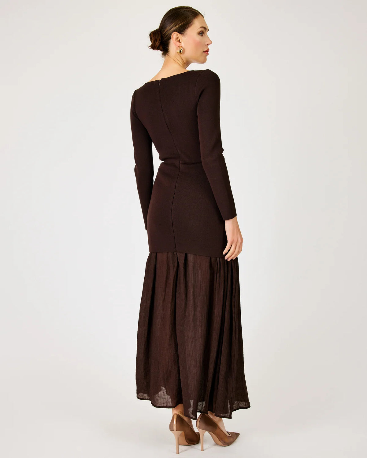 A woman modeling a dark brown long-sleeve maxi dress from the back, featuring a structured waist and a flowing pleated skirt, set against a plain light gray studio background.