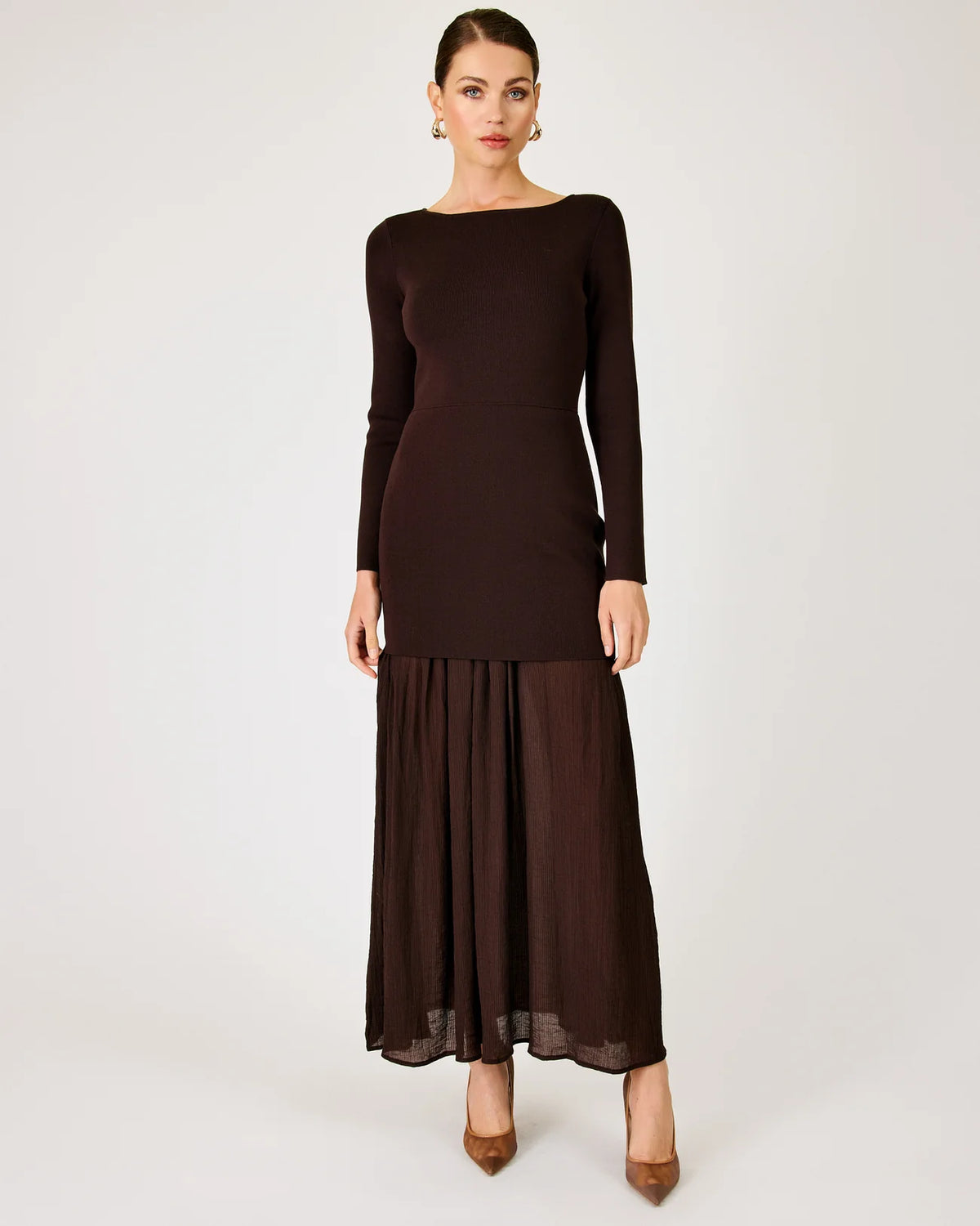 A woman modeling a dark brown long-sleeve maxi dress with a boat neckline and a pleated skirt, standing against a light gray studio background.