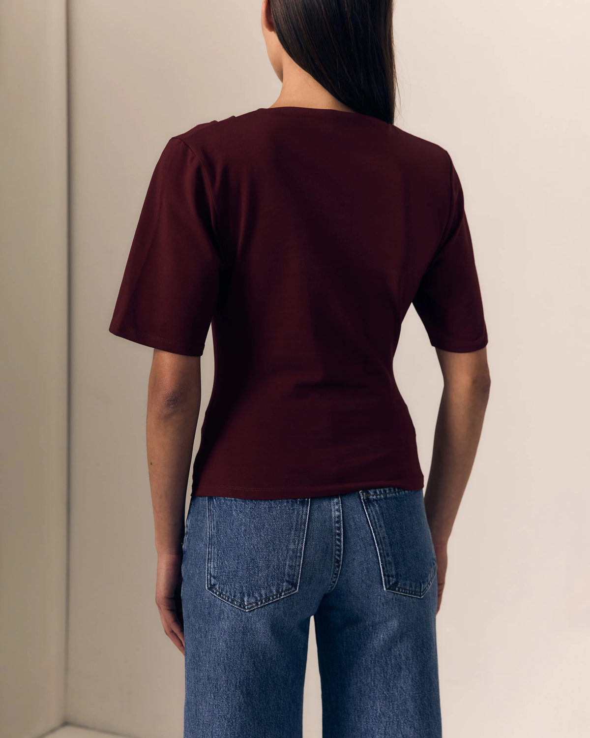 model wearing a short sleeve burgundy top with ruching across the shoulder and jeans on a cream background, back view 