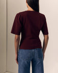 model wearing a short sleeve burgundy top with ruching across the shoulder and jeans on a cream background, back view 
