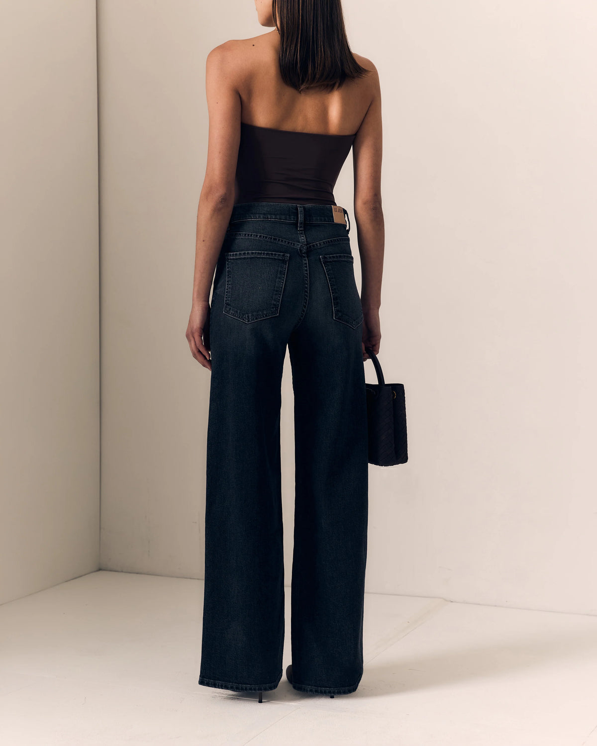 model wearing a strapless brown top with dark jeans on a cream background, back view 