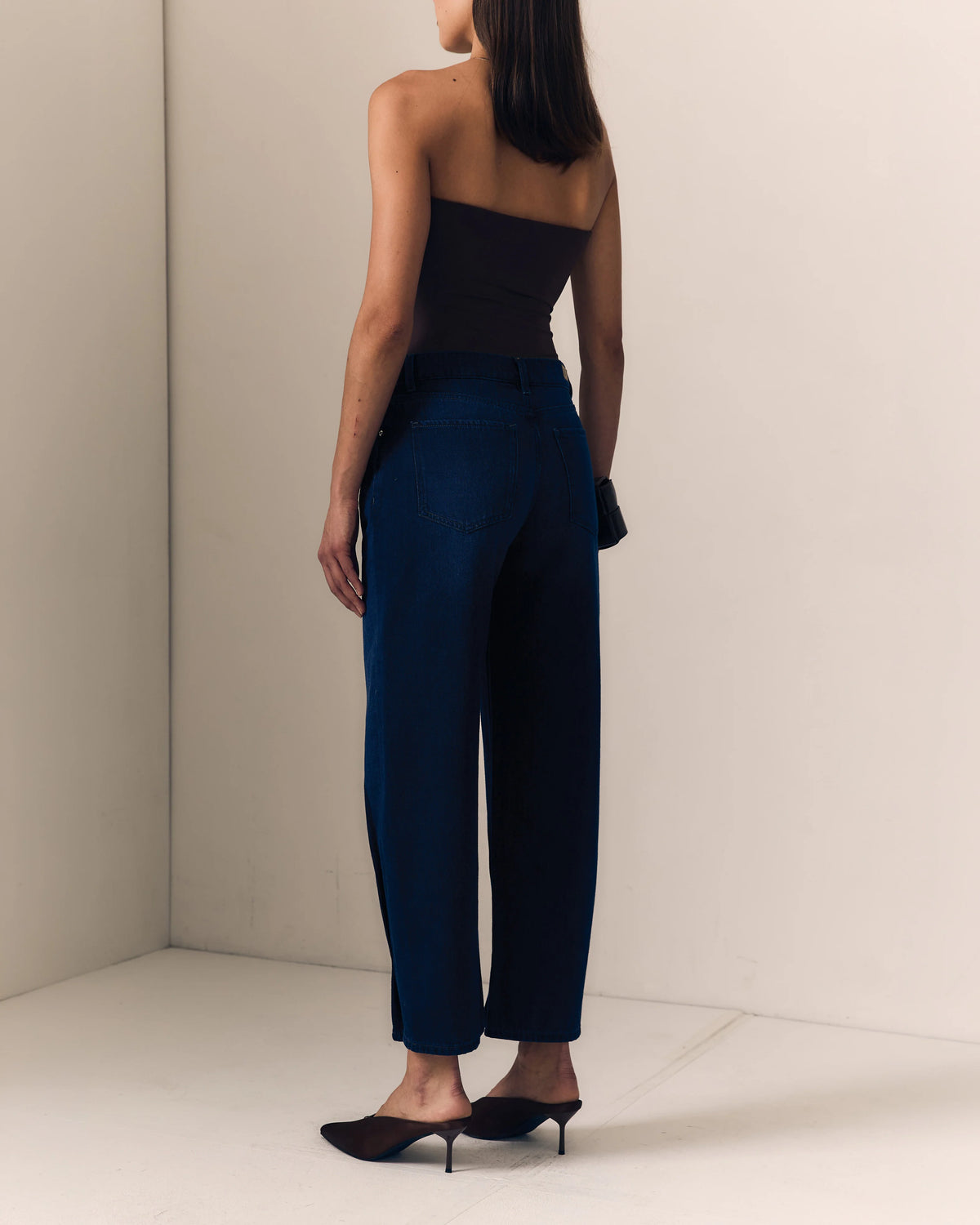model wearing dark blue jeans with a pinched hem and a strapless brown top on a cream background, back view