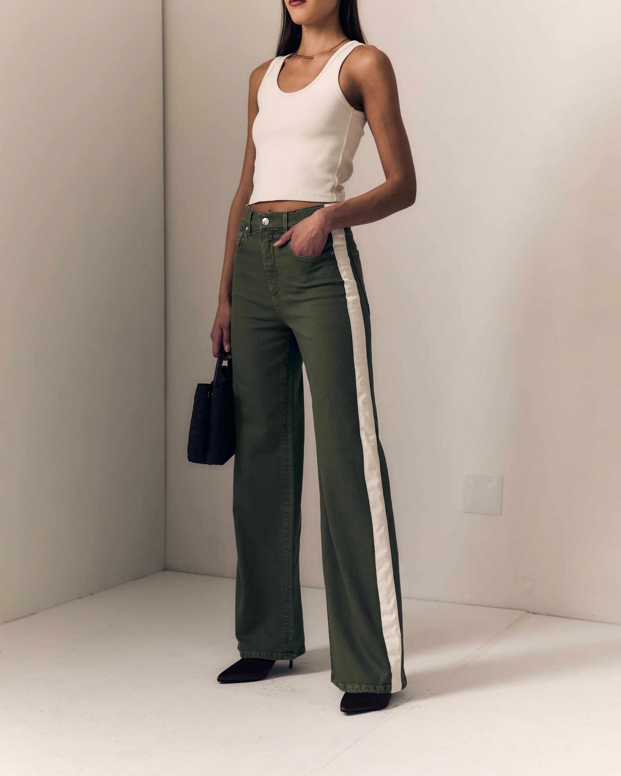 A model stands in a minimalist studio wearing olive green wide-leg pants with a white side stripe, paired with a cream tank top and black pointed shoes.