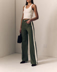 A model stands in a minimalist studio wearing olive green wide-leg pants with a white side stripe, paired with a cream tank top and black pointed shoes.